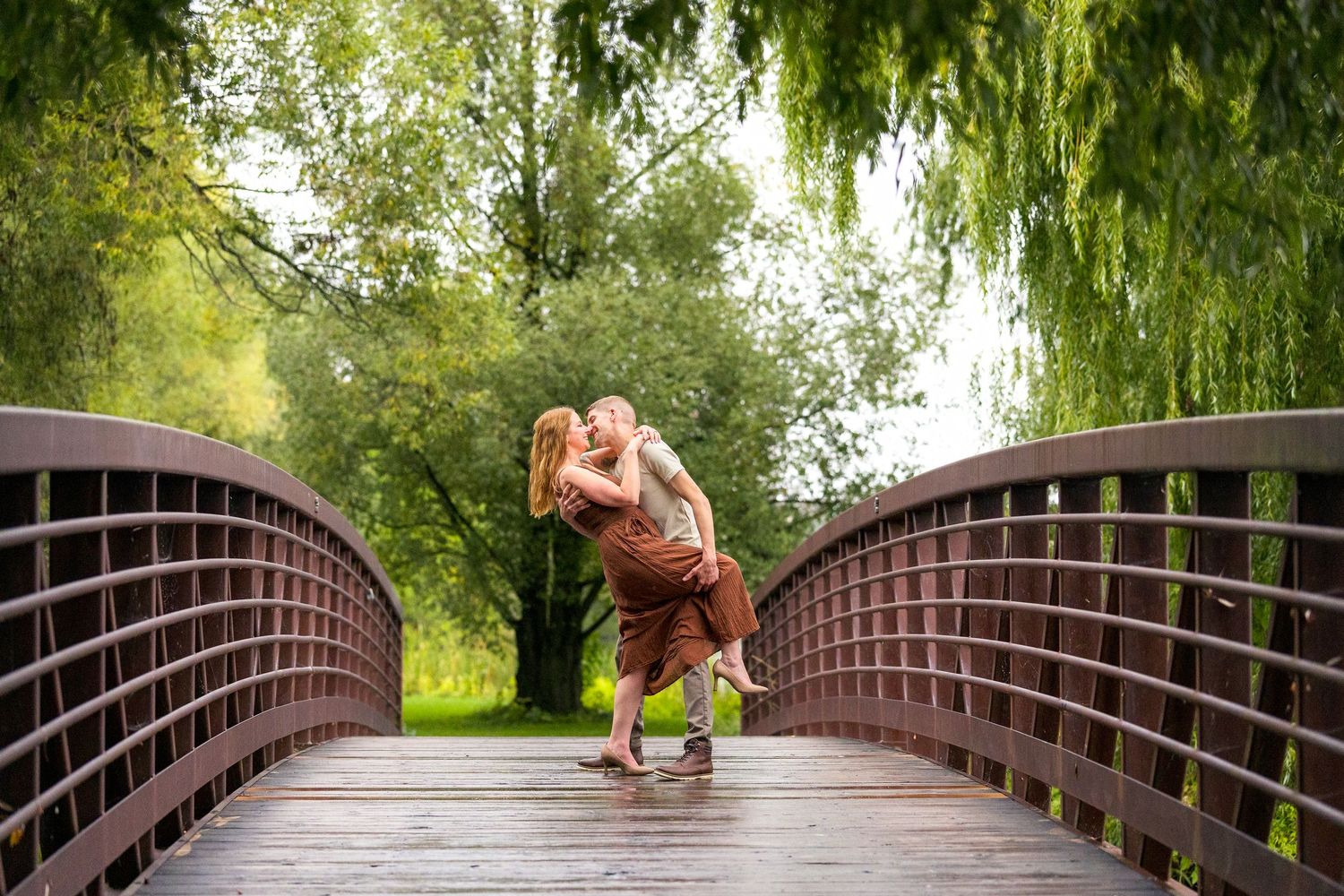 A couple shares a romantic kiss while dipping on a wooden footbridge surrounded by willow trees in Ottawa's Arboretum for their engagement
