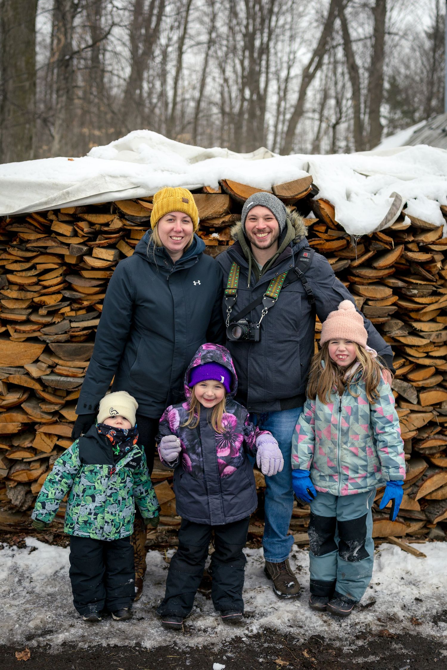 Ottawa wedding & portrait photographer with his family in winter attire against a backdrop of stacked firewood on a snowy day.