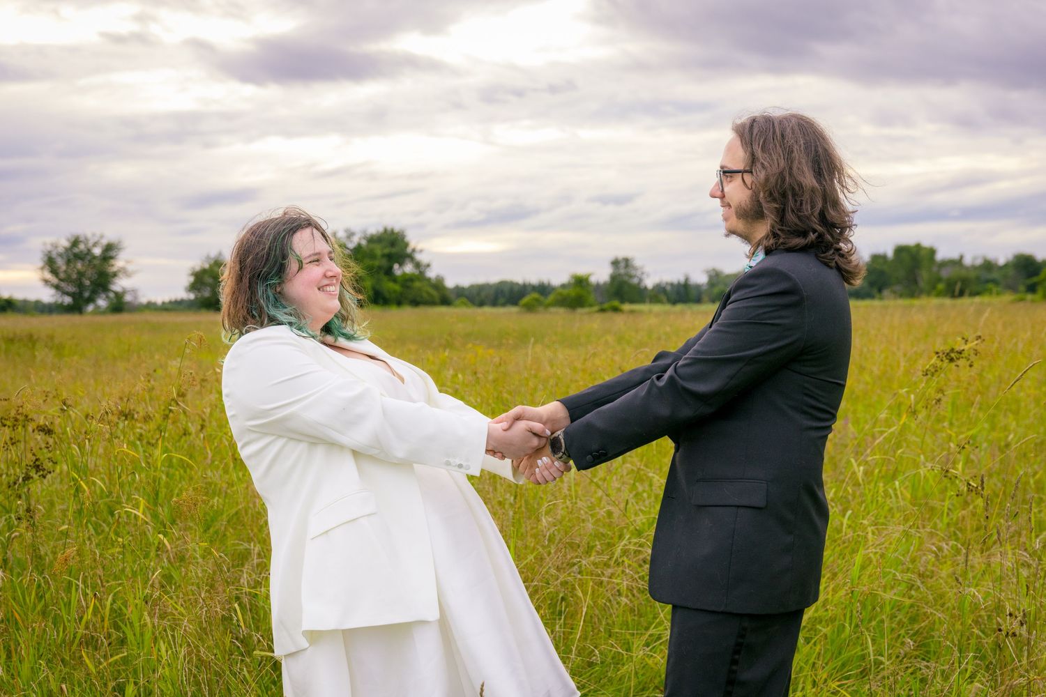 A couple in wedding attire holds hands and laughs together in a sunny meadow. Ottawa Wedding Photographer