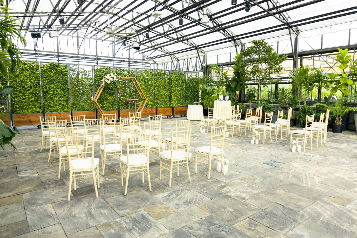 White chairs arranged in rows inside Aquatopia greenhouse venue with glass ceiling and lush greenery surrounding the space.