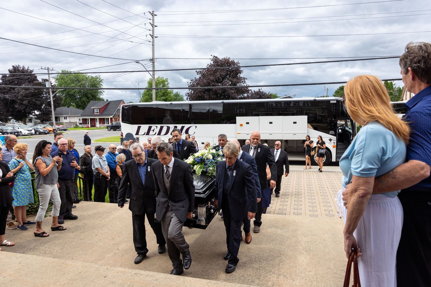 A large white tour bus arrives as people line up on both sides of a brick driveway outside a church for a celebration of life photographer