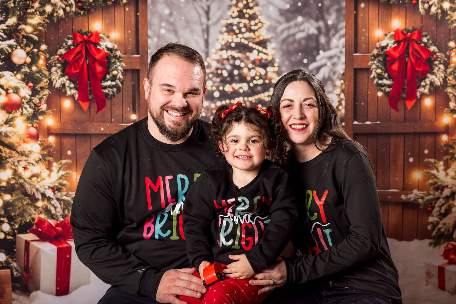 Family wearing matching Christmas sweaters poses in front of decorated trees and red bows in festive holiday setting.