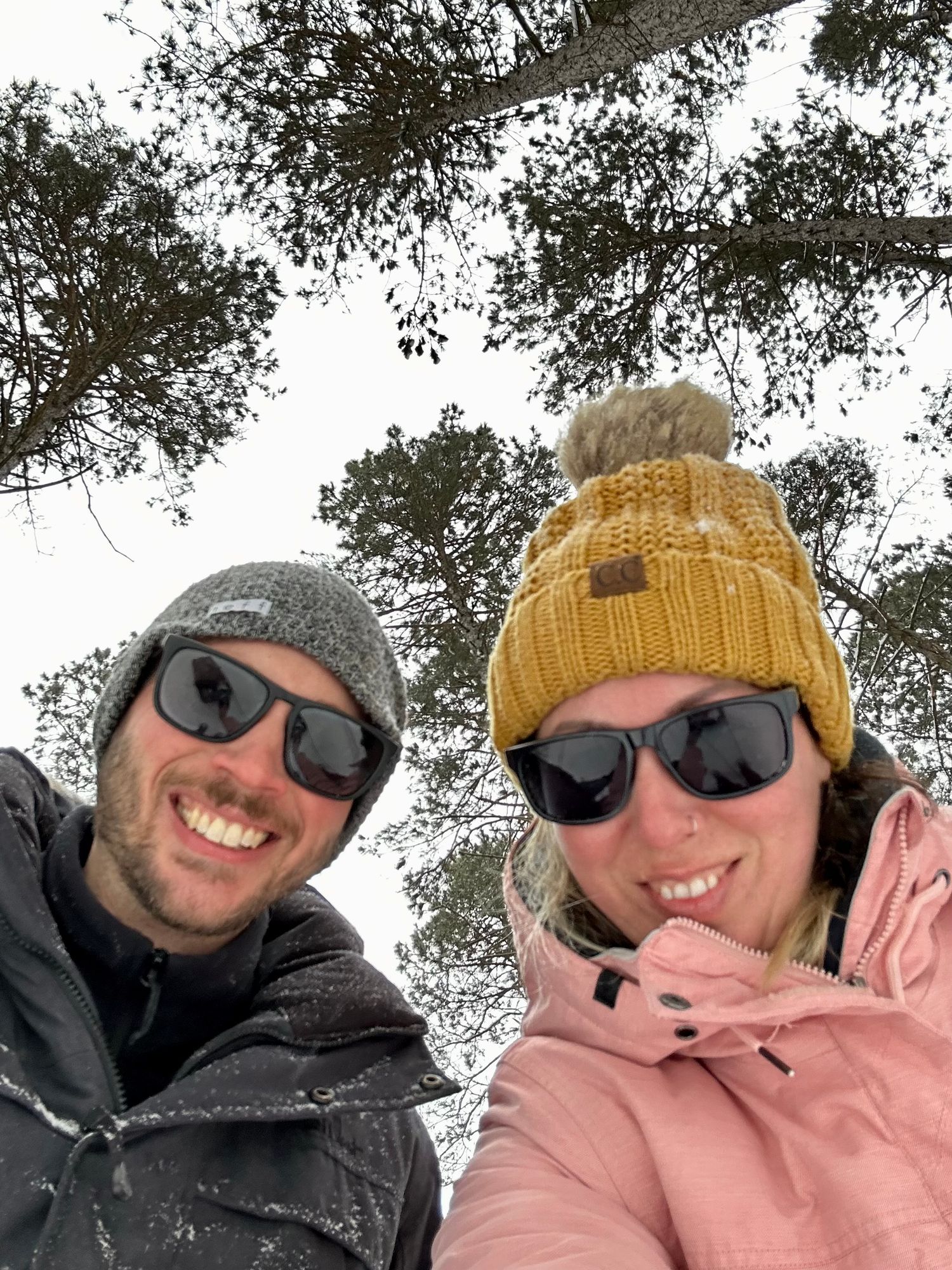 Ottawa wedding & lifestyle photographer & wife wearing winter hats and sunglasses smile together outdoors with trees in the background.
