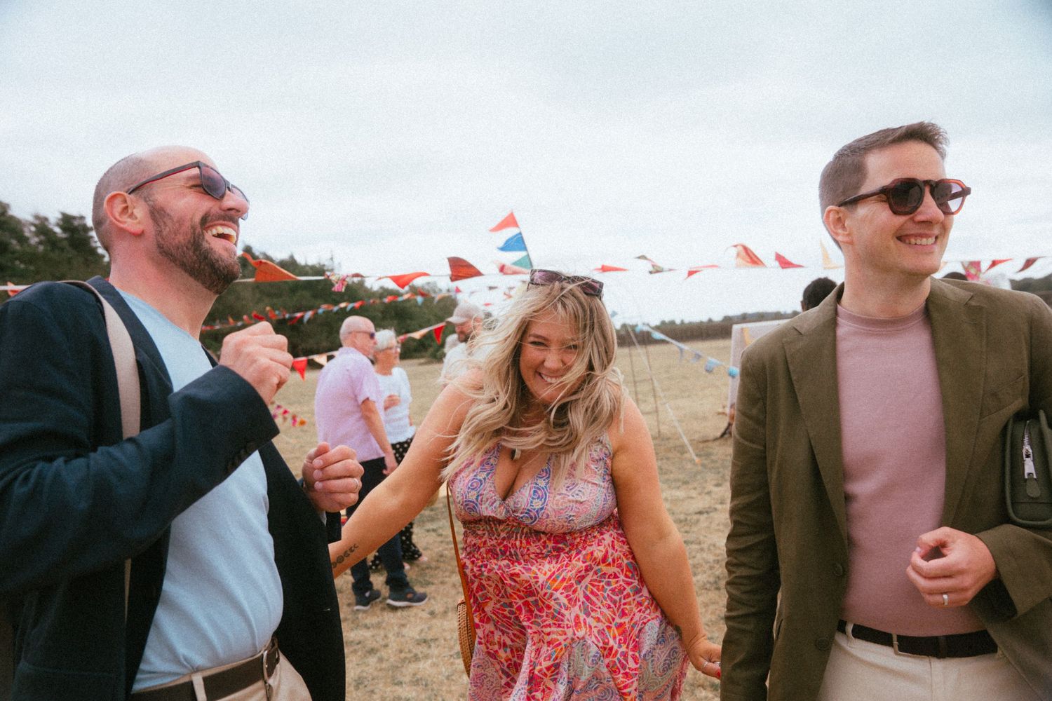 Three friends laughing and walking together at outdoor festival with colorful bunting flags.