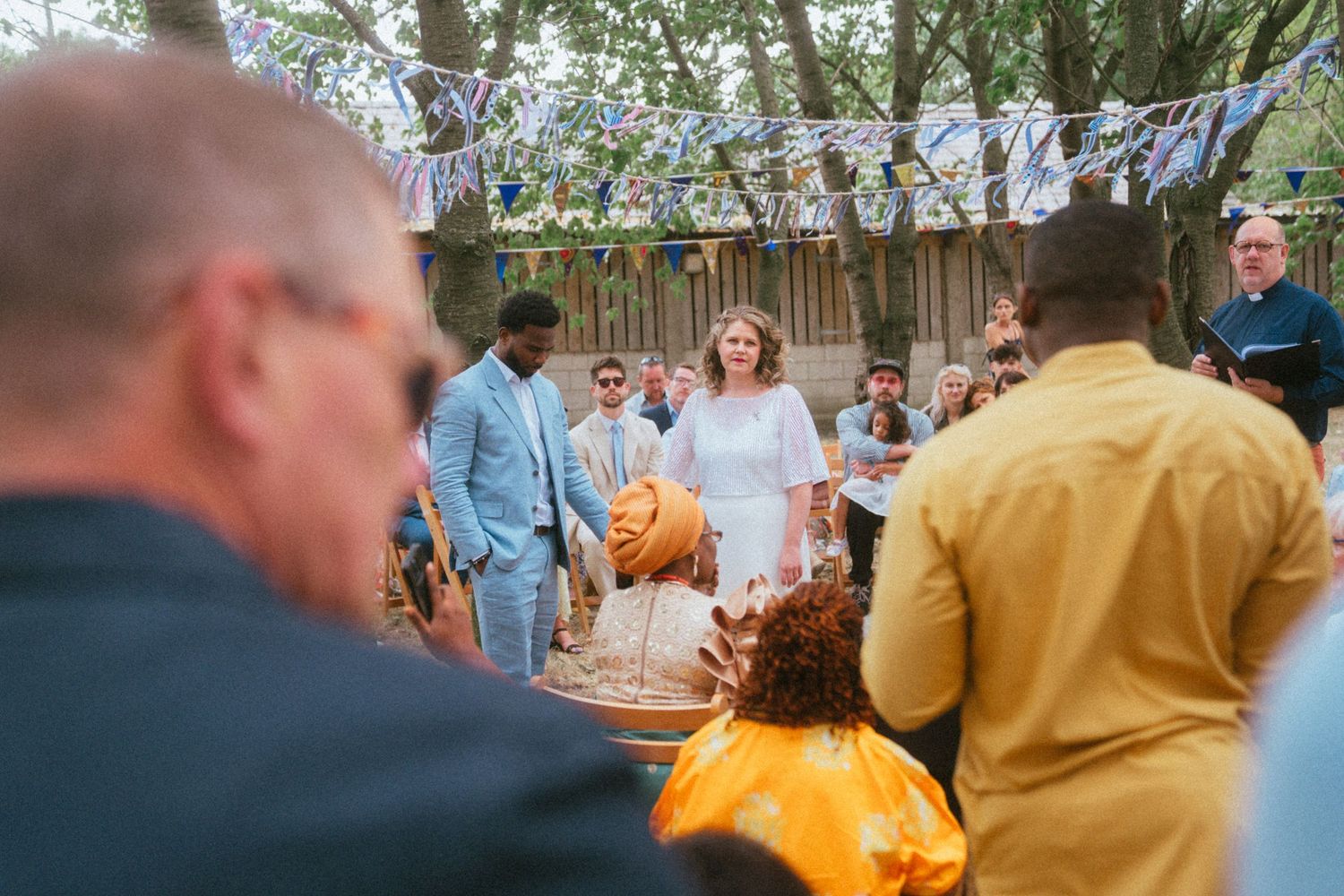 Bride in white dress stands at outdoor woodland wedding ceremony surrounded by guests in colorful attire.