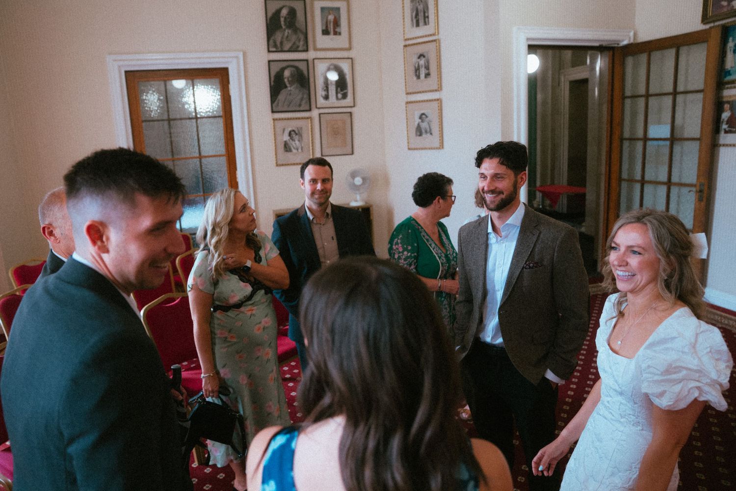 Wedding guests mingling and smiling in an elegant room decorated with framed photos on the walls.