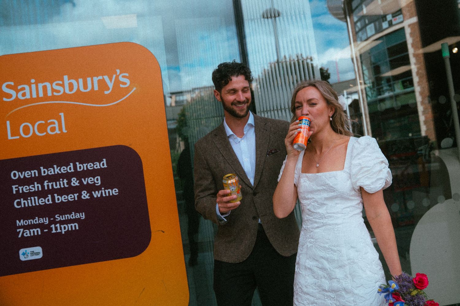 Bride and groom in wedding attire smile outside a Sainsbury's Local store holding drinks after their ceremony.