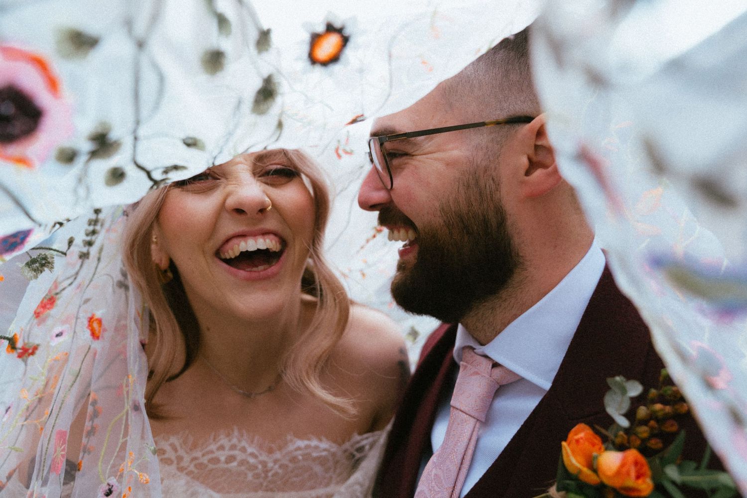 Joyful bride and groom laughing together under a floral embroidered veil on their wedding day.