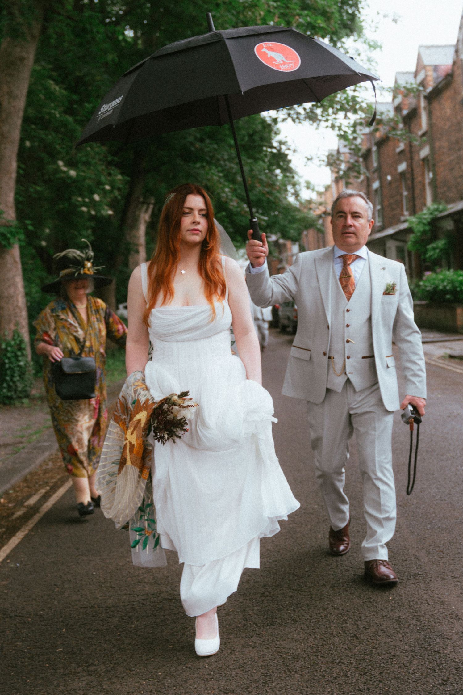 Bride in white dress walks with man in grey suit holding black umbrella on tree-lined street.
