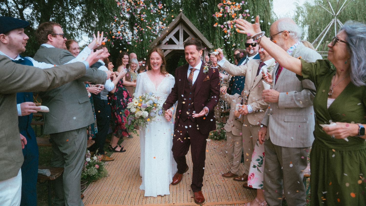 Bride and groom walk through confetti shower with cheering wedding guests outside a rustic chapel.