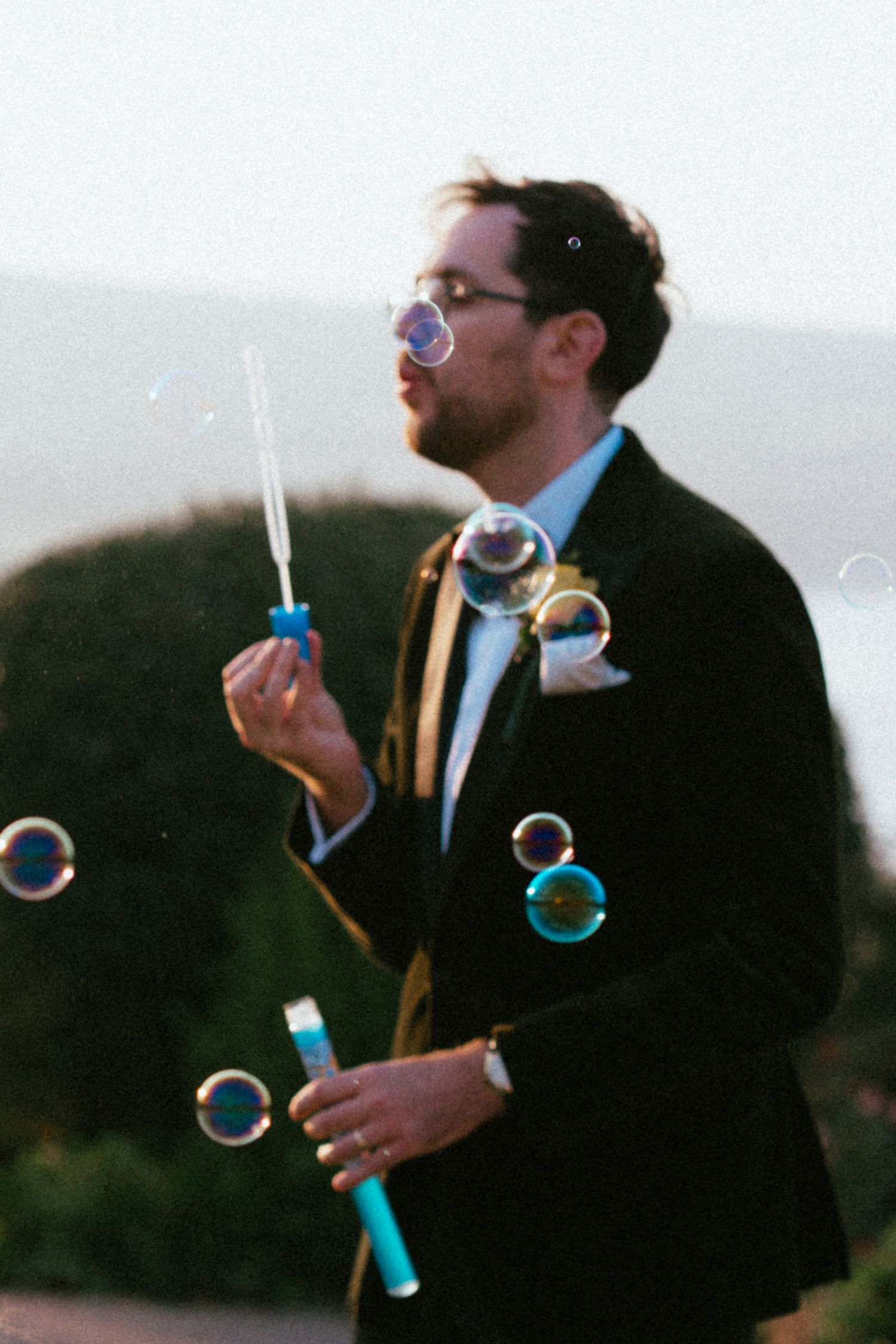 Groom in a dark suit and bow tie blowing soap bubbles with a blue wand at an outdoor wedding celebration at sunset.