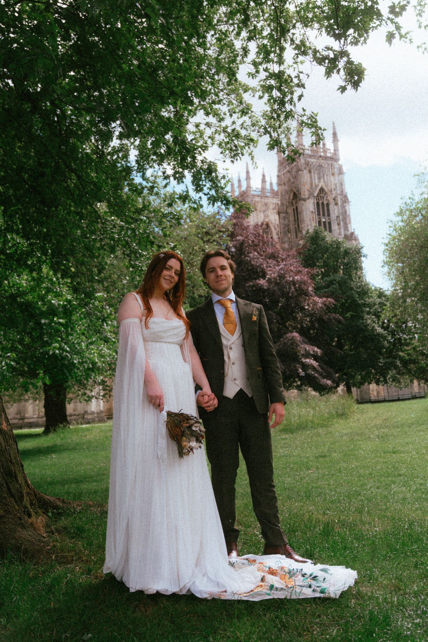 Wedding couple holding hands under tree with Gothic cathedral tower in background.