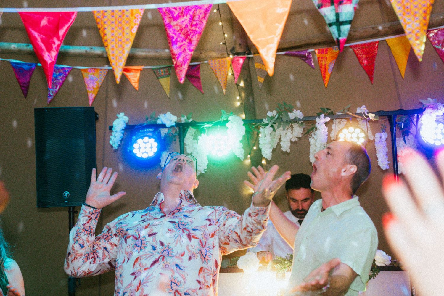 Guests dancing joyfully at colorful wedding reception with vibrant bunting and festive party lights overhead.