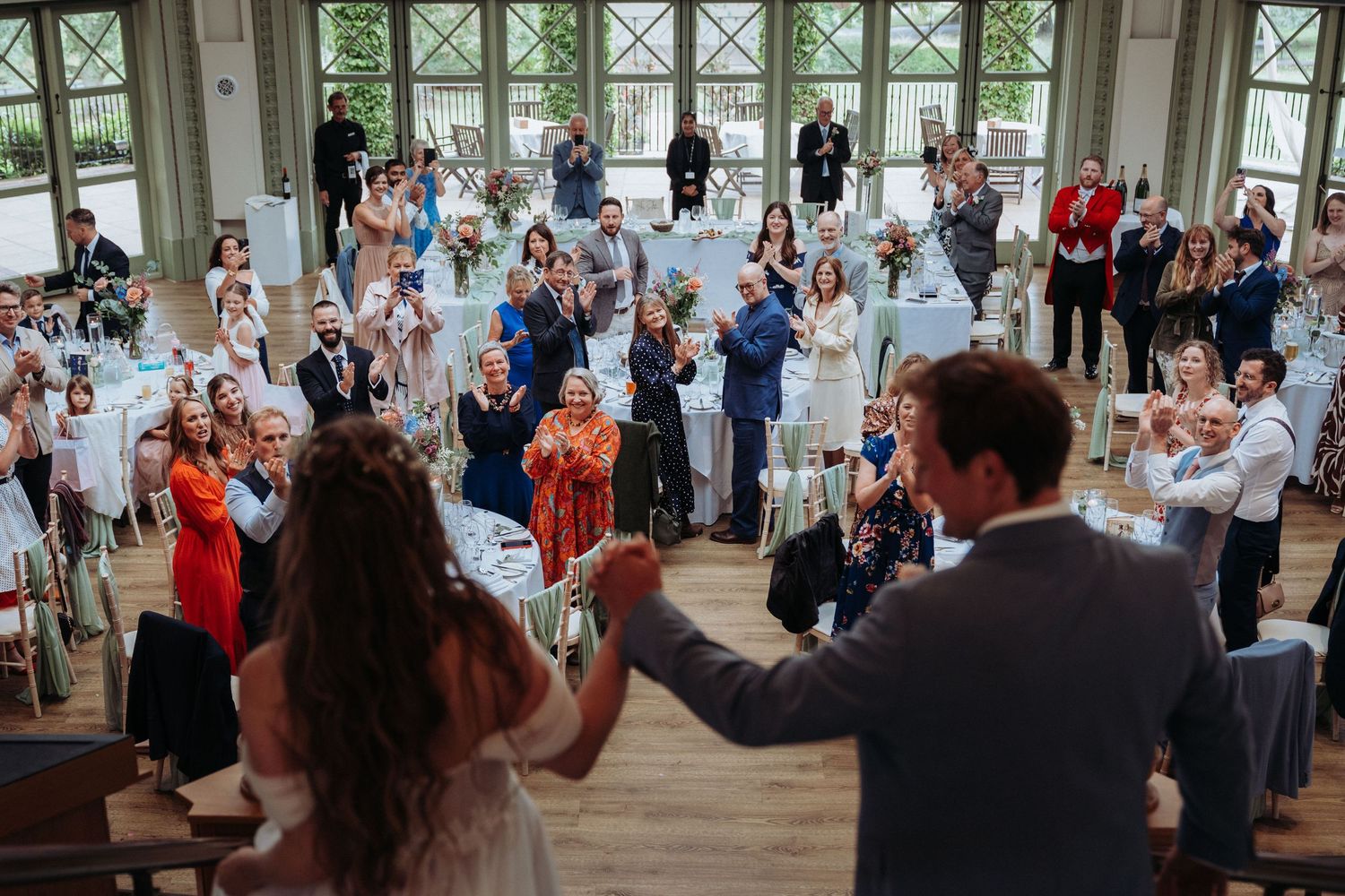 Bride and groom enter reception hall as guests clap and cheer in a bright glass conservatory.