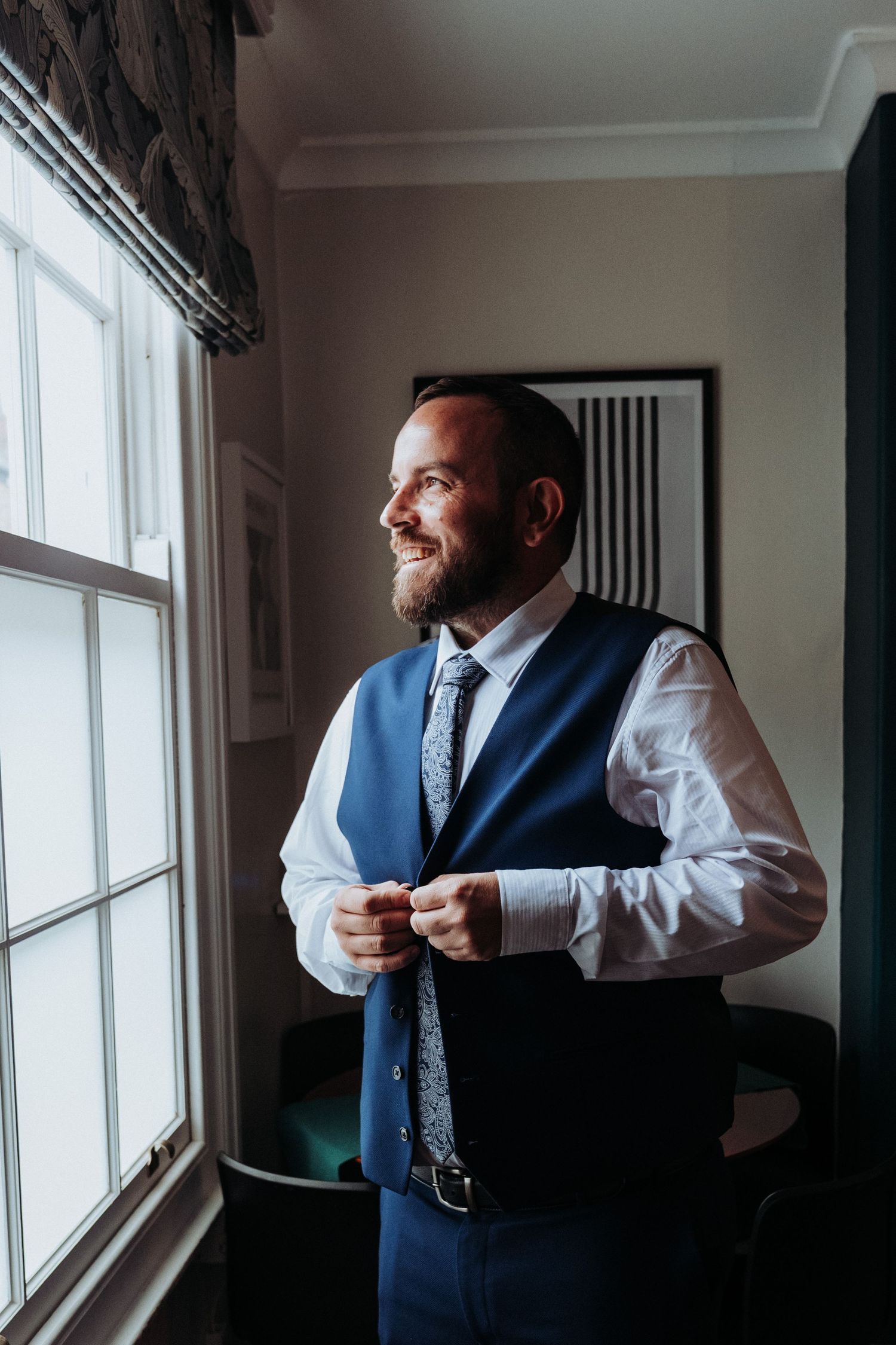 Smiling bearded groom in blue vest and tie buttoning his waistcoat while standing by a bright window.