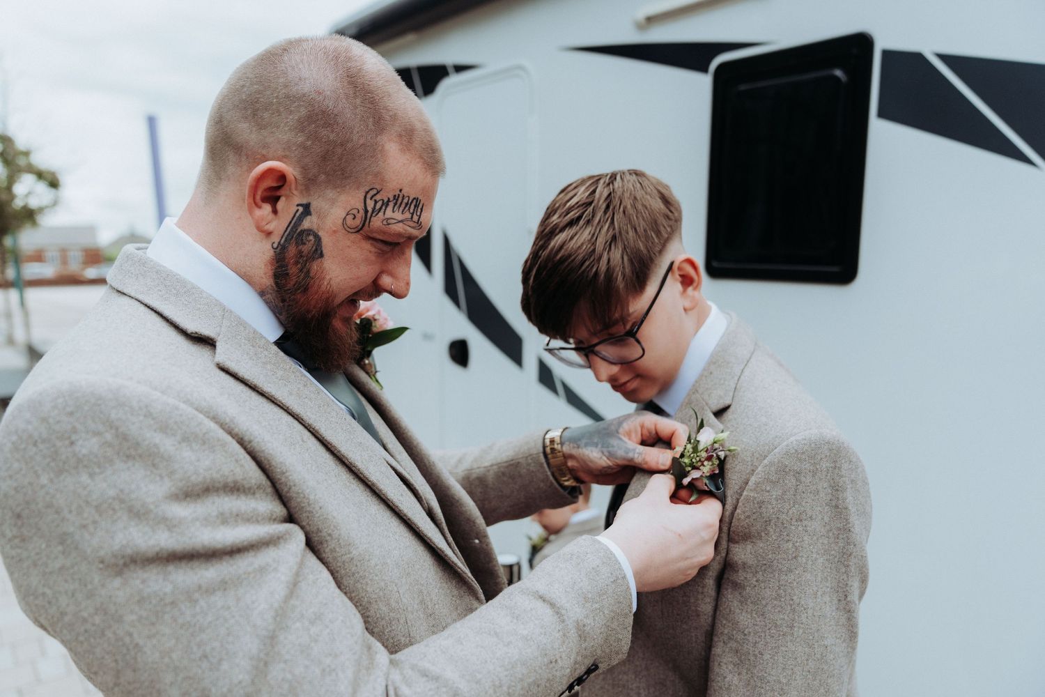 Tattooed man in grey suit pinning a boutonniere on a young boy in matching suit outdoors.