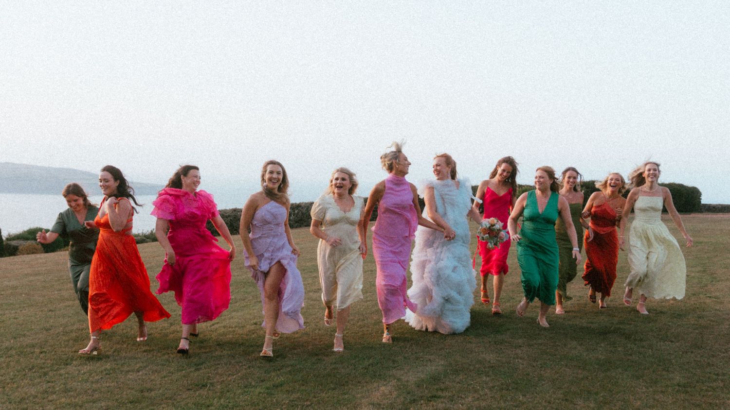 Group of women in colorful dresses running together across a grassy hilltop overlooking the sea on a bright, hazy day.