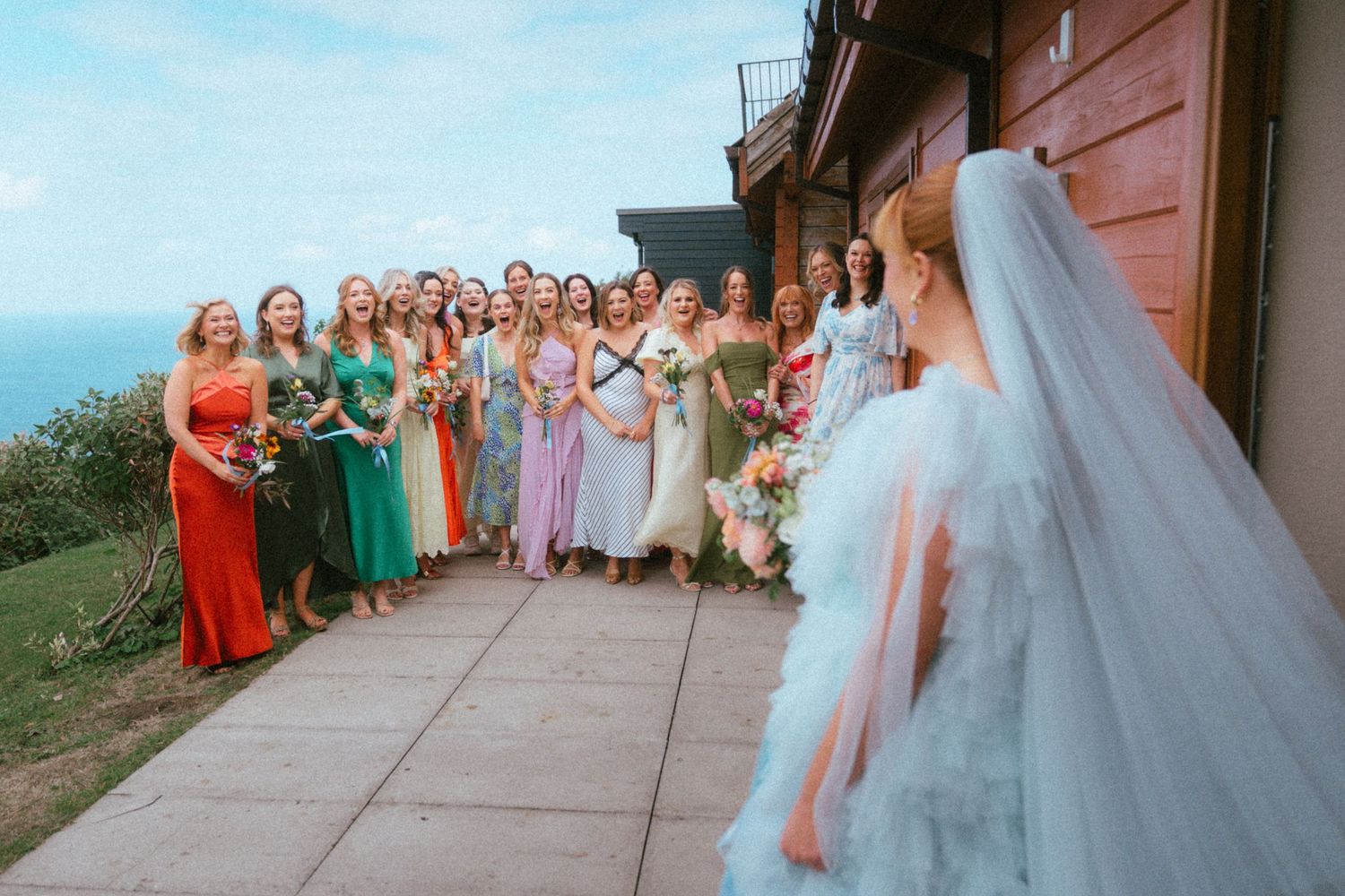 Bride in a flowing white tulle gown faces a group of bridesmaids in colorful dresses holding bouquets on a terrace overlooking the ocean.