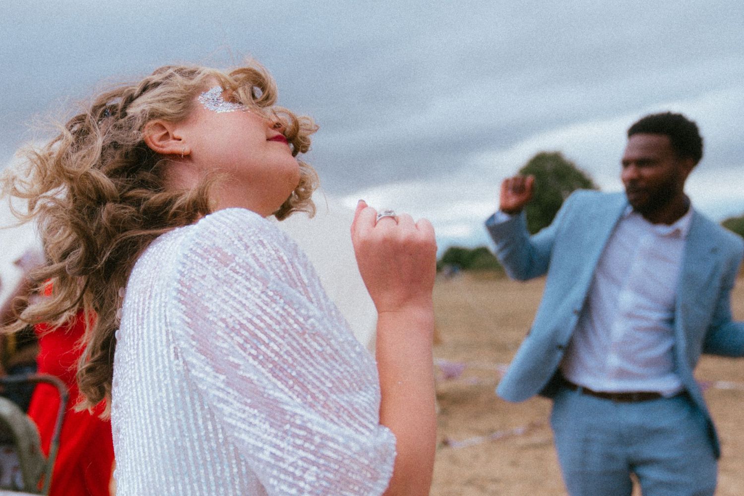 A curly-haired woman in a white sequin top laughs joyfully while dancing outdoors at a celebration.