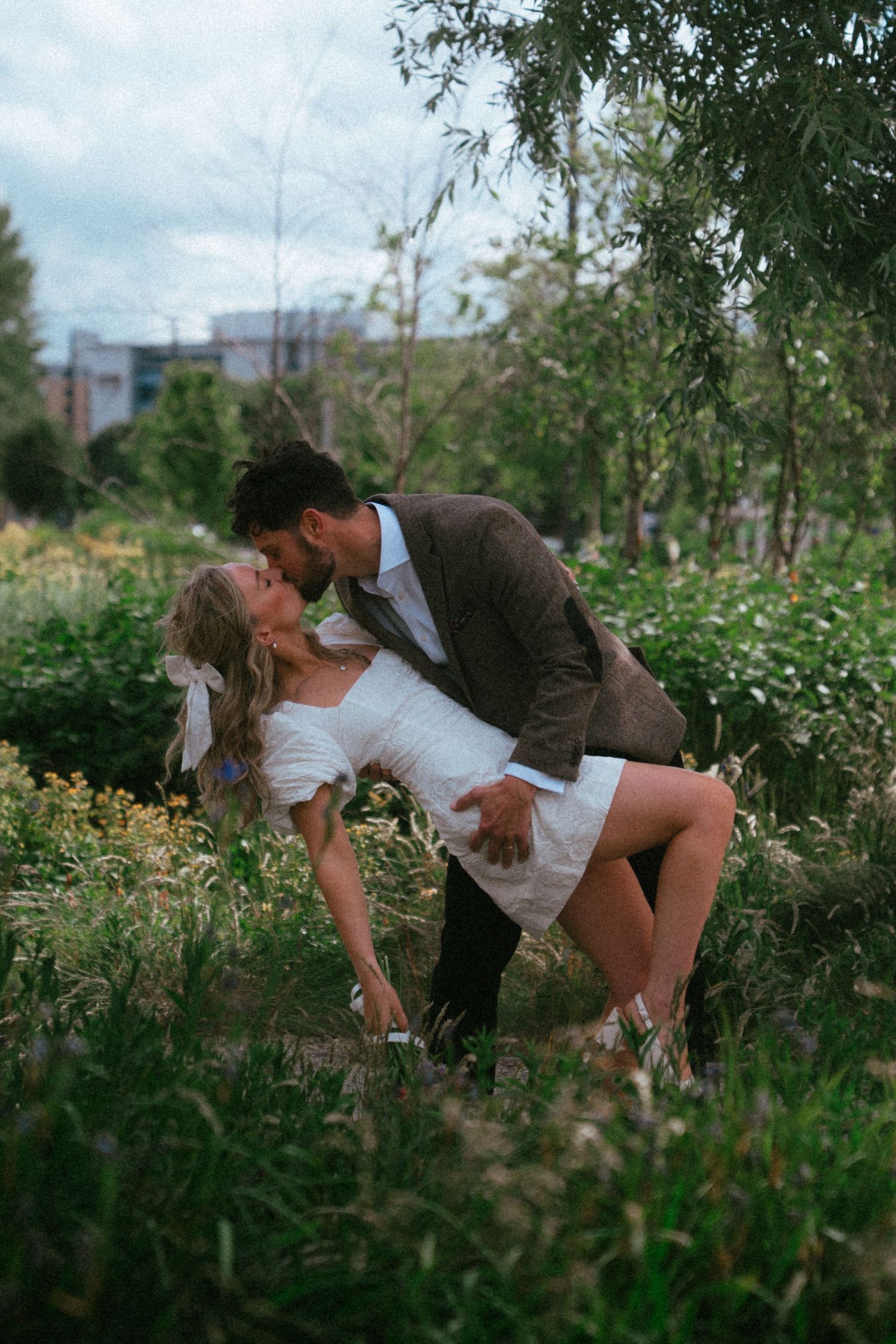 A couple shares a romantic dip kiss surrounded by lush greenery in an outdoor garden setting.