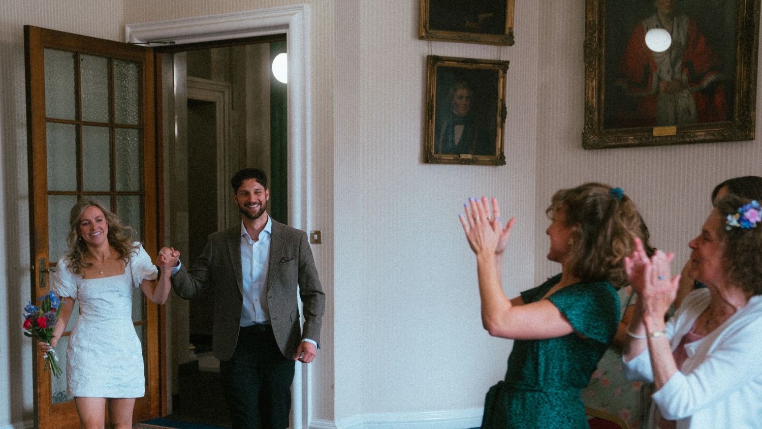 Newlywed couple entering room greeted by applauding guests at an elegant indoor wedding celebration.