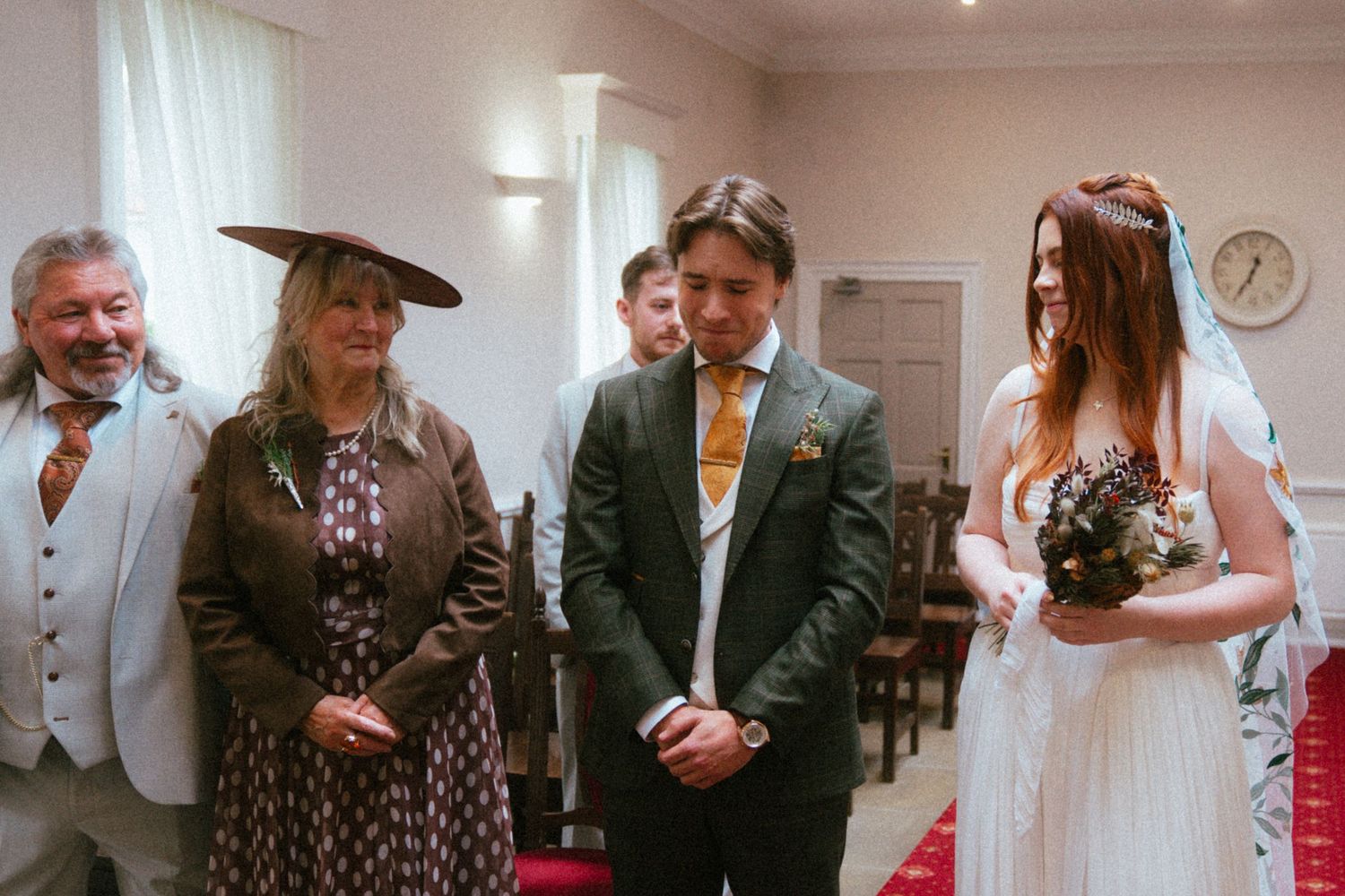 Bride and groom stand at wedding ceremony with family members, bride holding bouquet in white dress with veil.