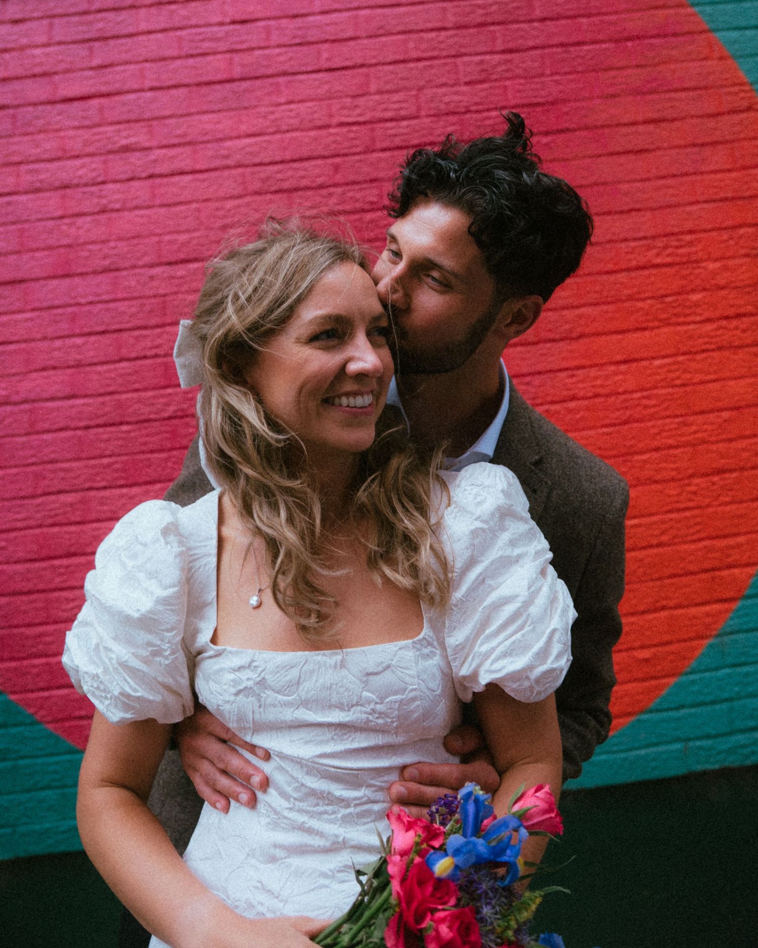 A groom kisses his bride on the cheek as she smiles, holding a bouquet, against a colorful red and green mural wall.