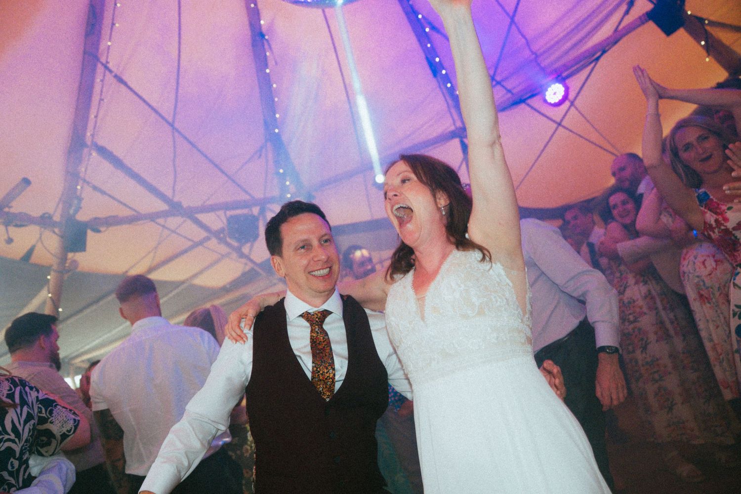 Bride and groom dancing joyfully together under colorful lights inside a wedding reception tent.