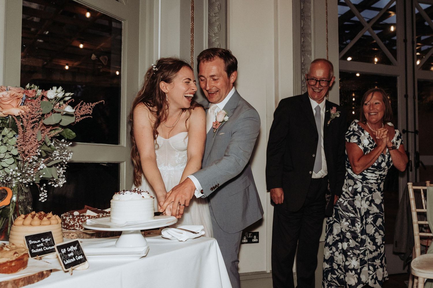 Smiling bride and groom cutting white wedding cake together while guests applaud in elegant venue.