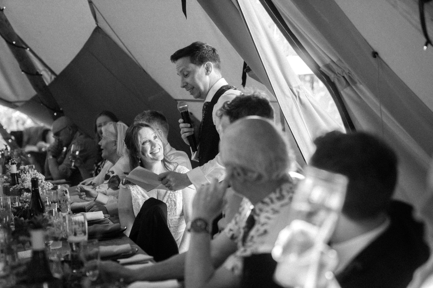 Black and white wedding reception photo of guests laughing at a long table inside a marquee tent.