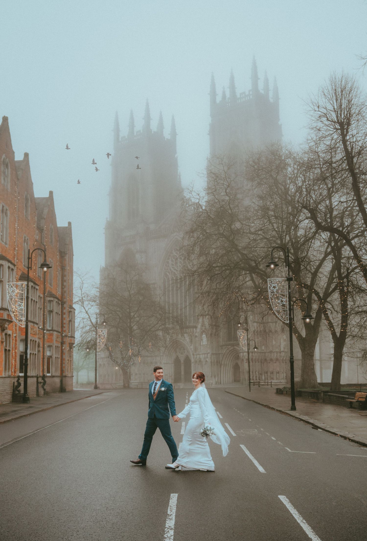Newlyweds walk hand in hand across foggy street with majestic Gothic cathedral in background.