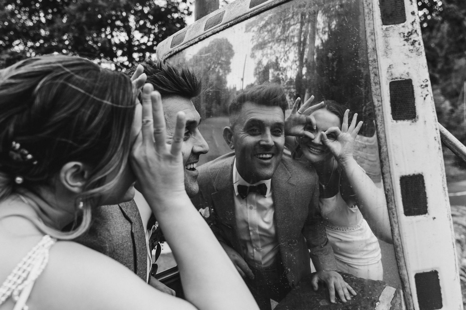 Black and white photo of wedding guests laughing and making funny faces reflected in a vintage vehicle mirror.