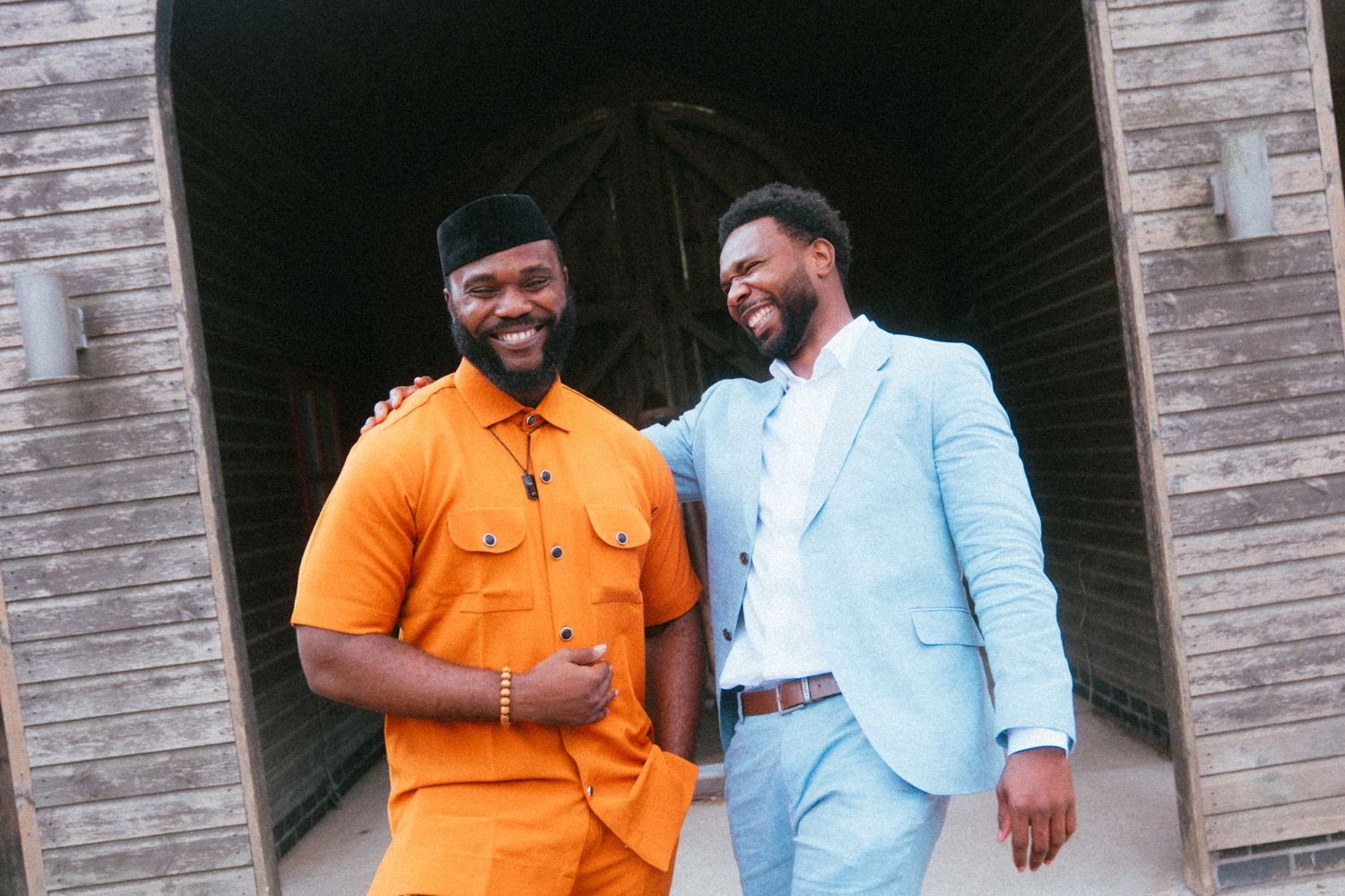 Two men in vibrant attire laugh together outside a rustic barn, one in orange and one in light blue suit.