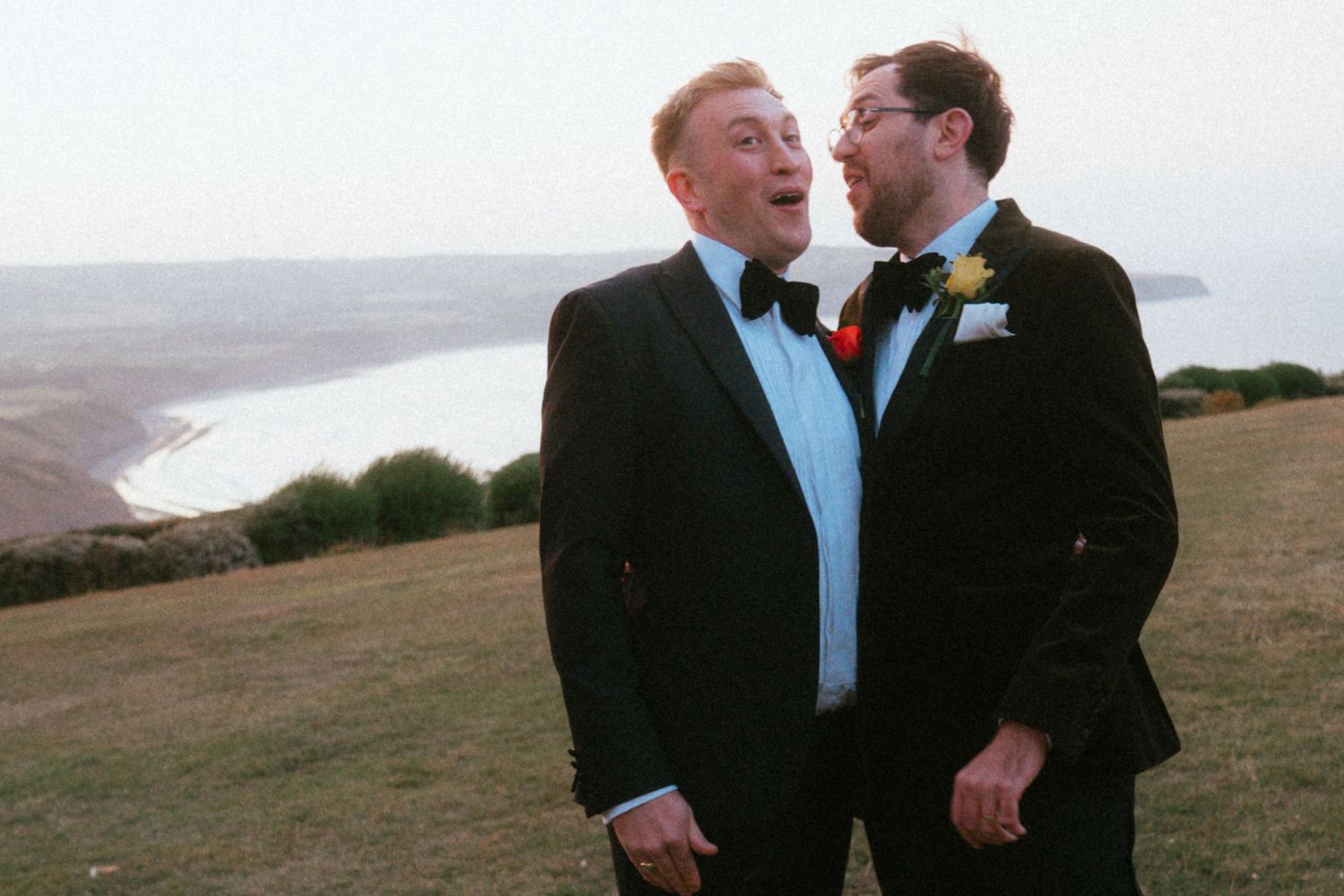 Two men in black tuxedos with bow ties and boutonnieres laugh together on a grassy clifftop overlooking the sea at a wedding.