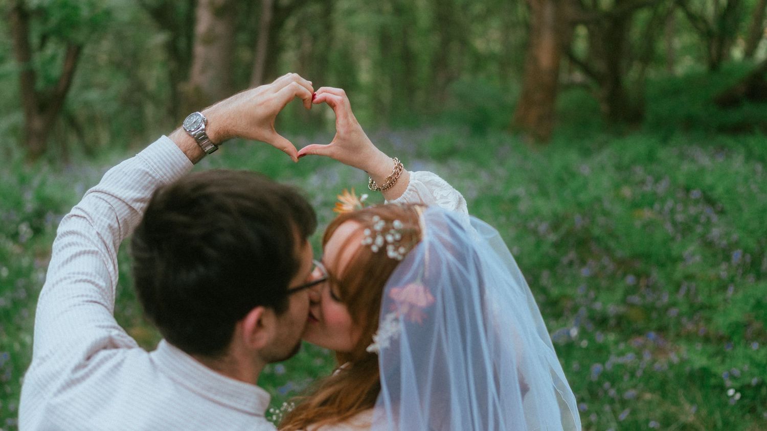 Bride and groom kiss in a sunlit forest while forming a heart shape with their hands above their heads.