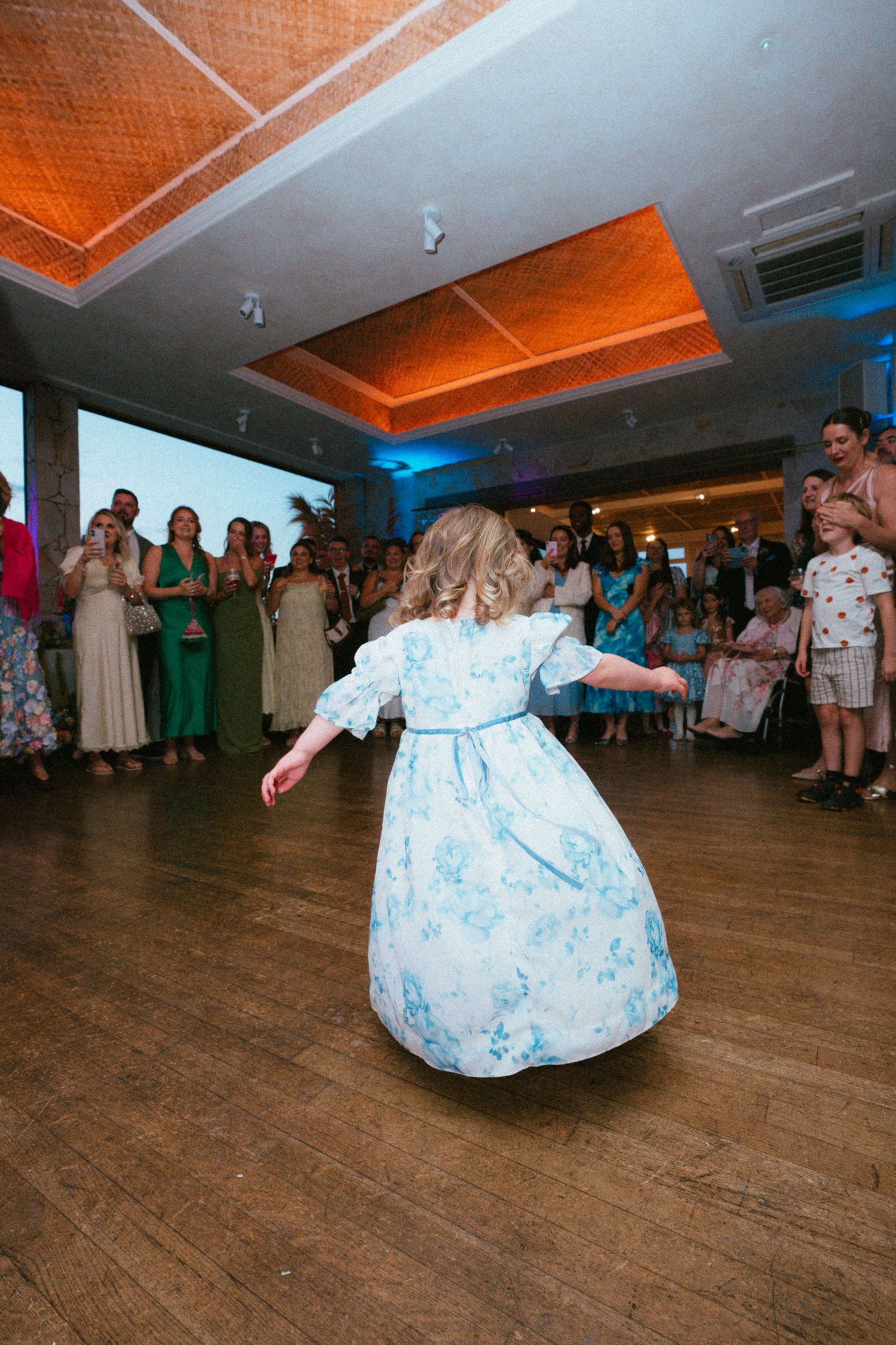 A young girl in a blue floral dress twirls on a wooden dance floor as wedding guests watch from the edge of the reception.