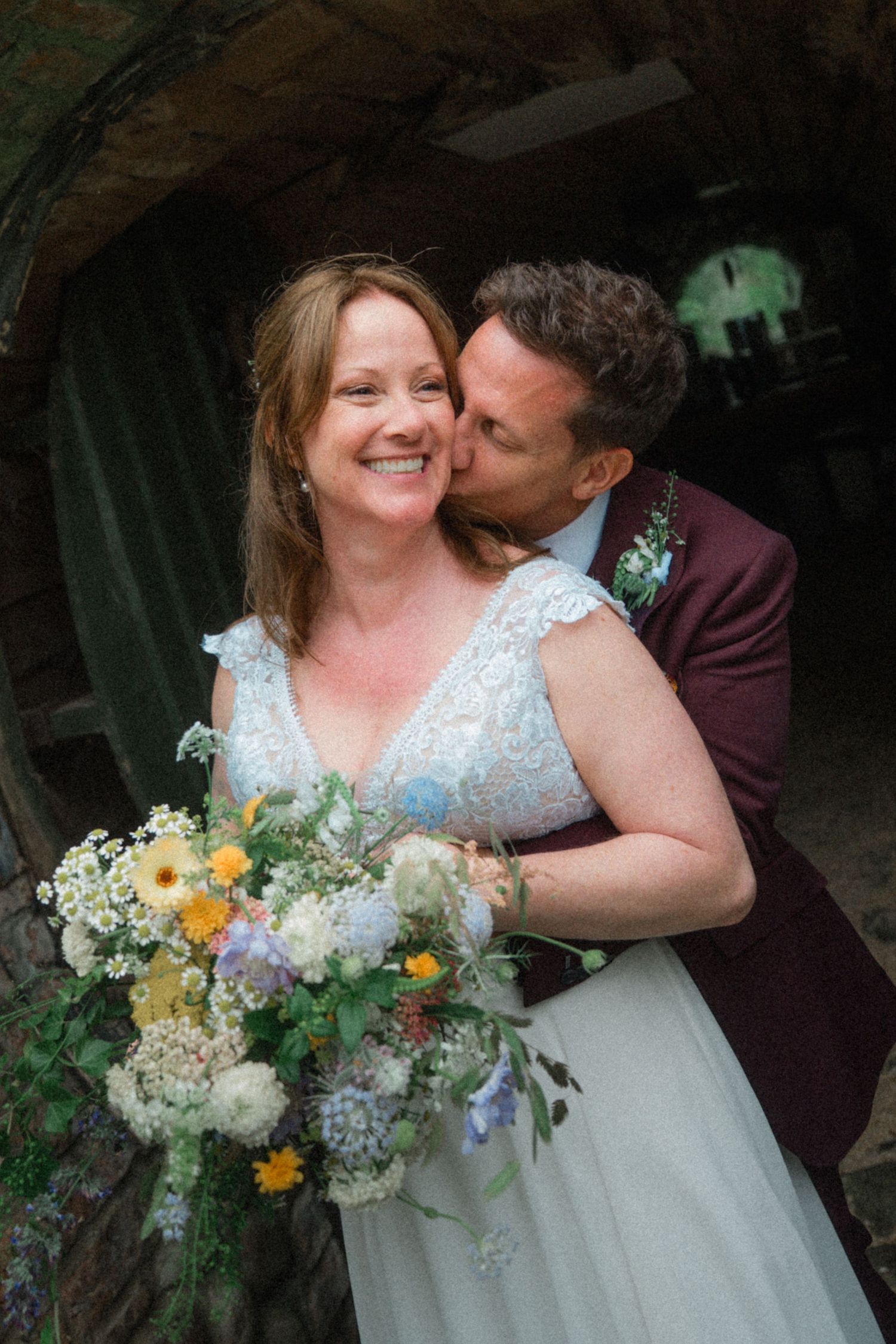 Groom in burgundy suit kisses bride on cheek as she smiles holding a wildflower bouquet.