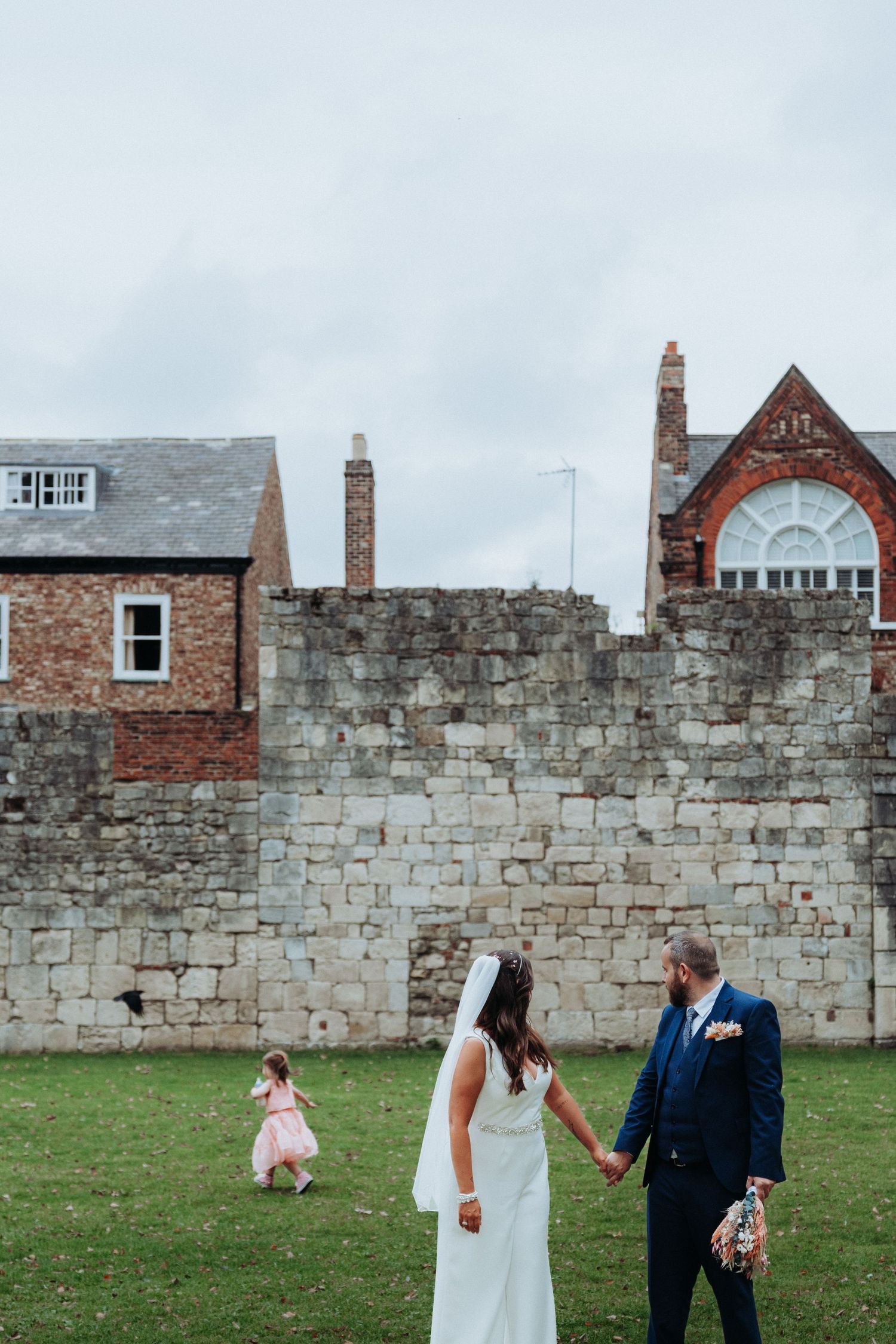 Bride and groom hold hands watching flower girl run across lawn beside historic stone walls in York.