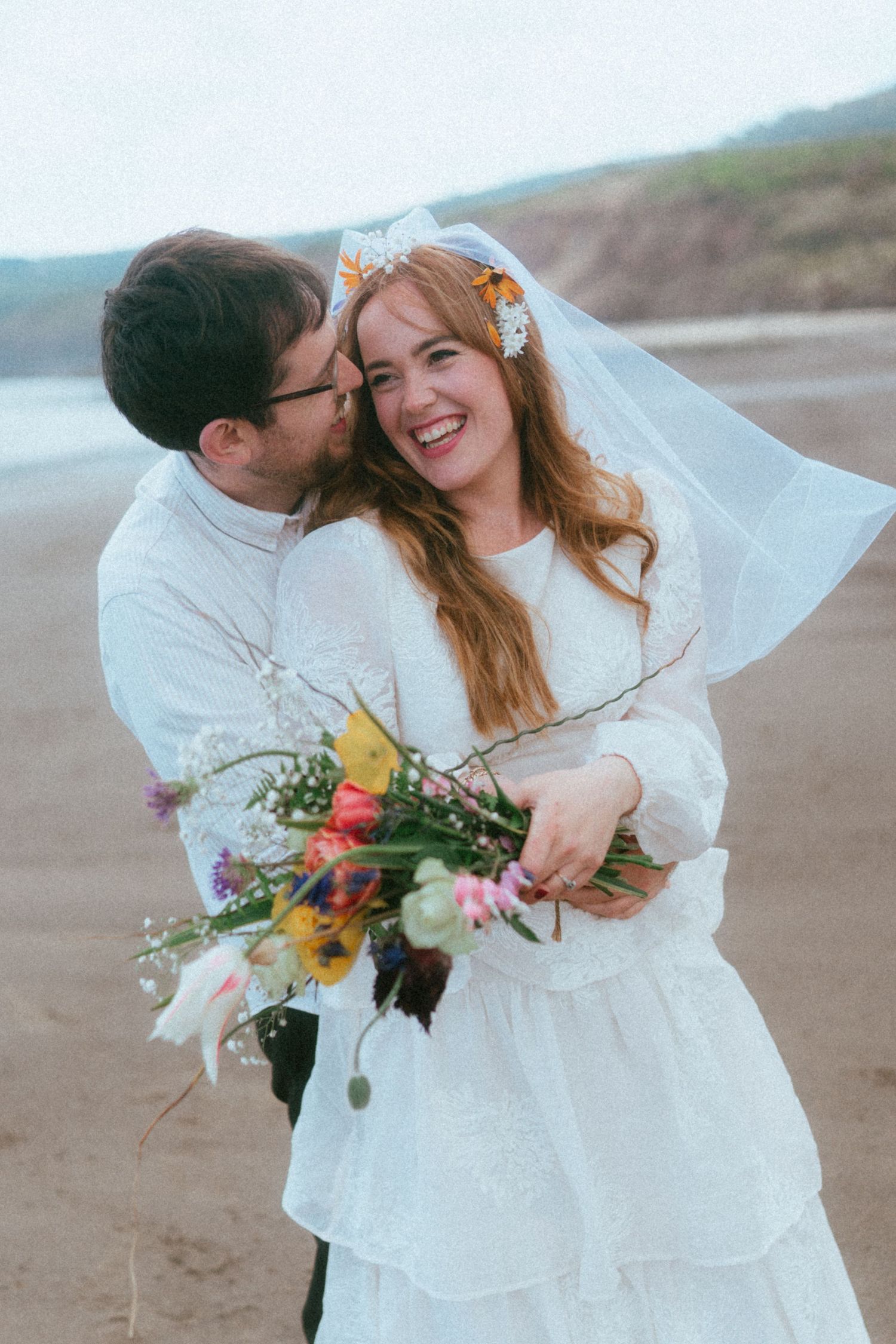 Couple embracing on beach in wedding attire, bride in white dress with veil holding colorful wildflower bouquet, smiling joyfully.