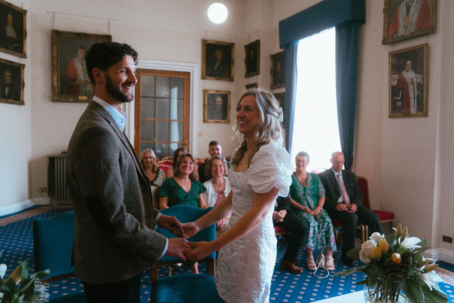 Bride and groom exchange vows holding hands at a civil wedding ceremony with guests seated in a blue-carpeted room.