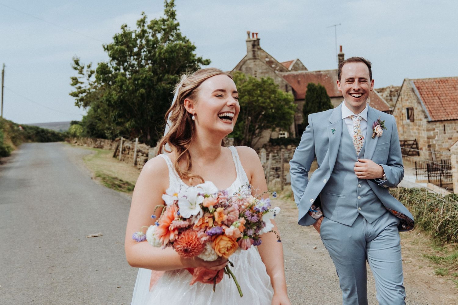 Laughing bride holding colorful bouquet walks with groom in blue suit along a country road.