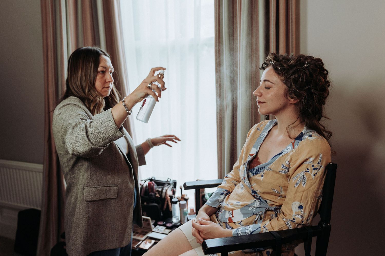 Professional makeup artist applying hairspray to bride during wedding morning preparation indoors.