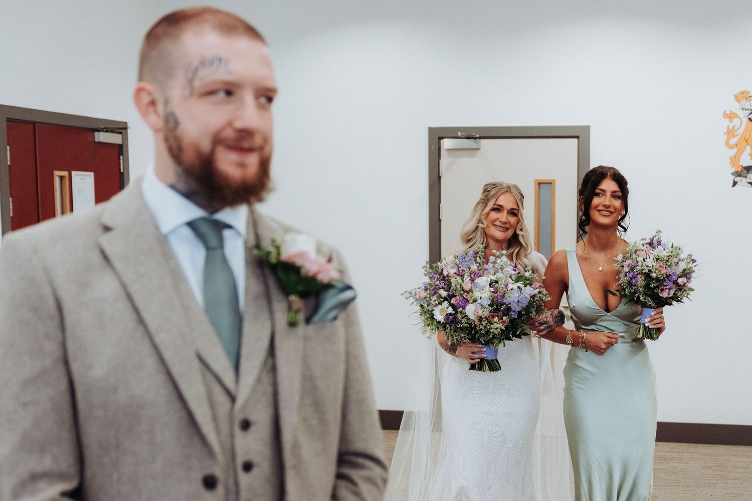Tattooed groom in grey suit stands as bridesmaids in mint dresses smile behind him holding bouquets.