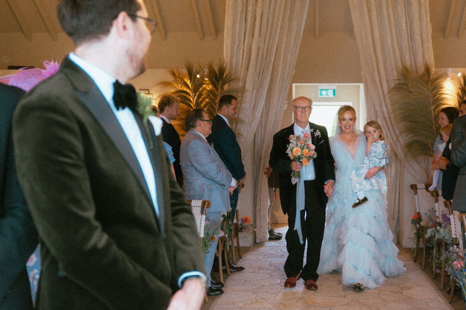 Bride in a flowing blue gown walks down the aisle with an older man and a small child as wedding guests look on.