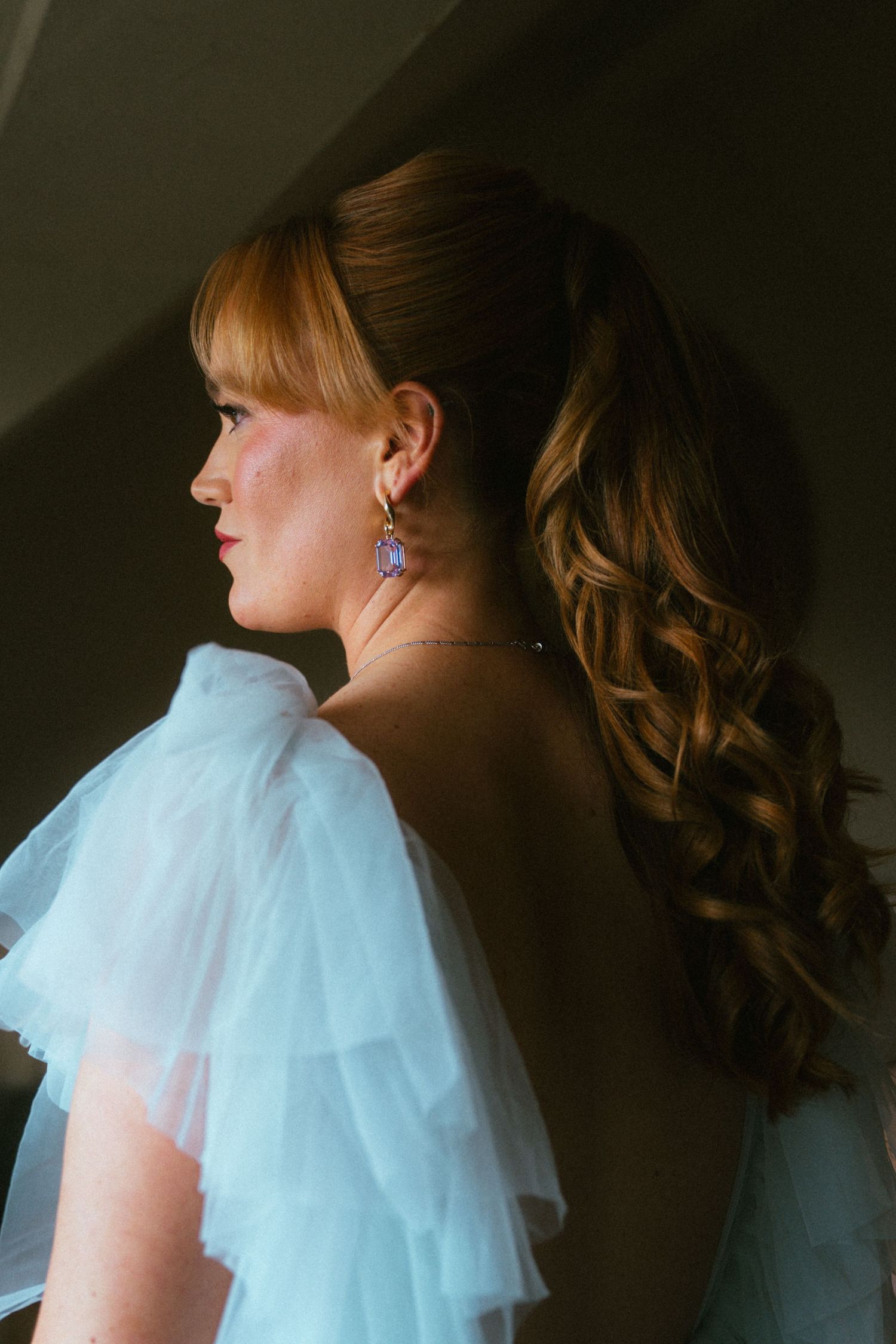 Bride in profile wearing a light blue tulle ruffled gown and drop earrings, her auburn curls styled in a high ponytail.