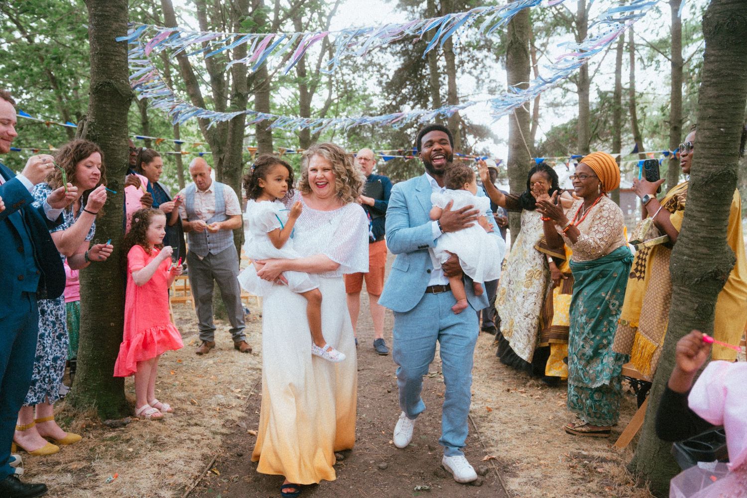 Newlyweds walking with children through cheering guests at outdoor woodland wedding celebration.