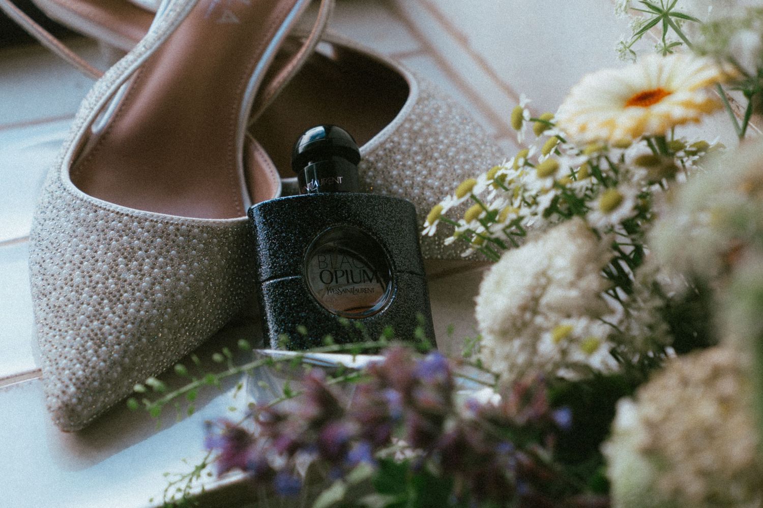 Black Opium YSL perfume bottle beside sparkly wedding heels and wildflowers.