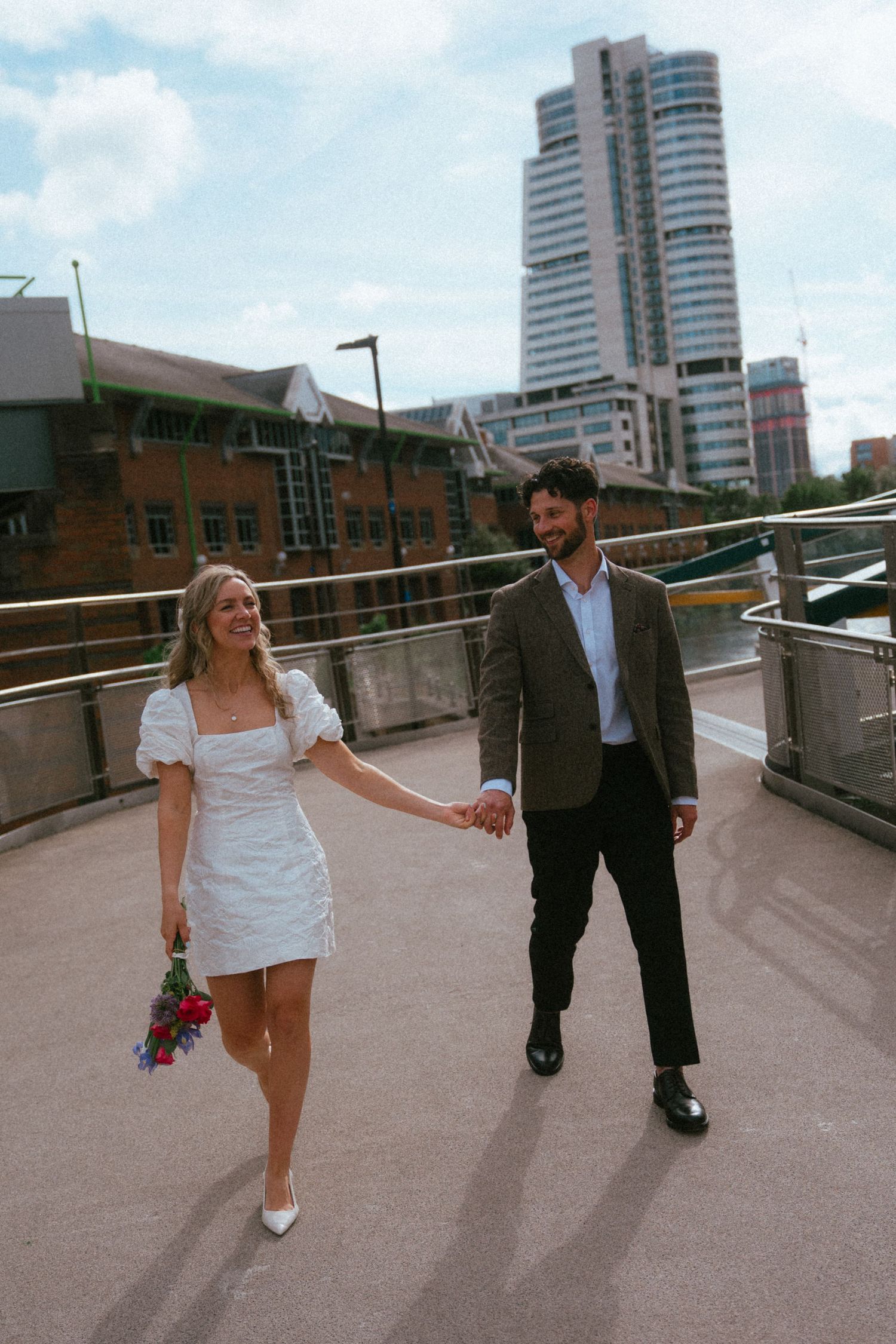 Couple holding hands on a bridge in white dress and suit with city skyline behind them.