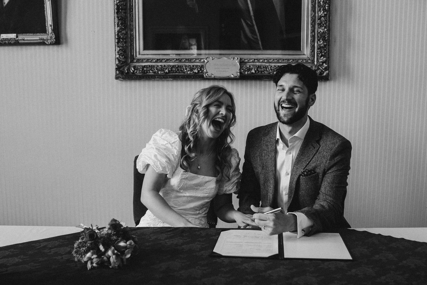 Black and white photo of a laughing bride and groom signing their wedding register at a ceremony table.