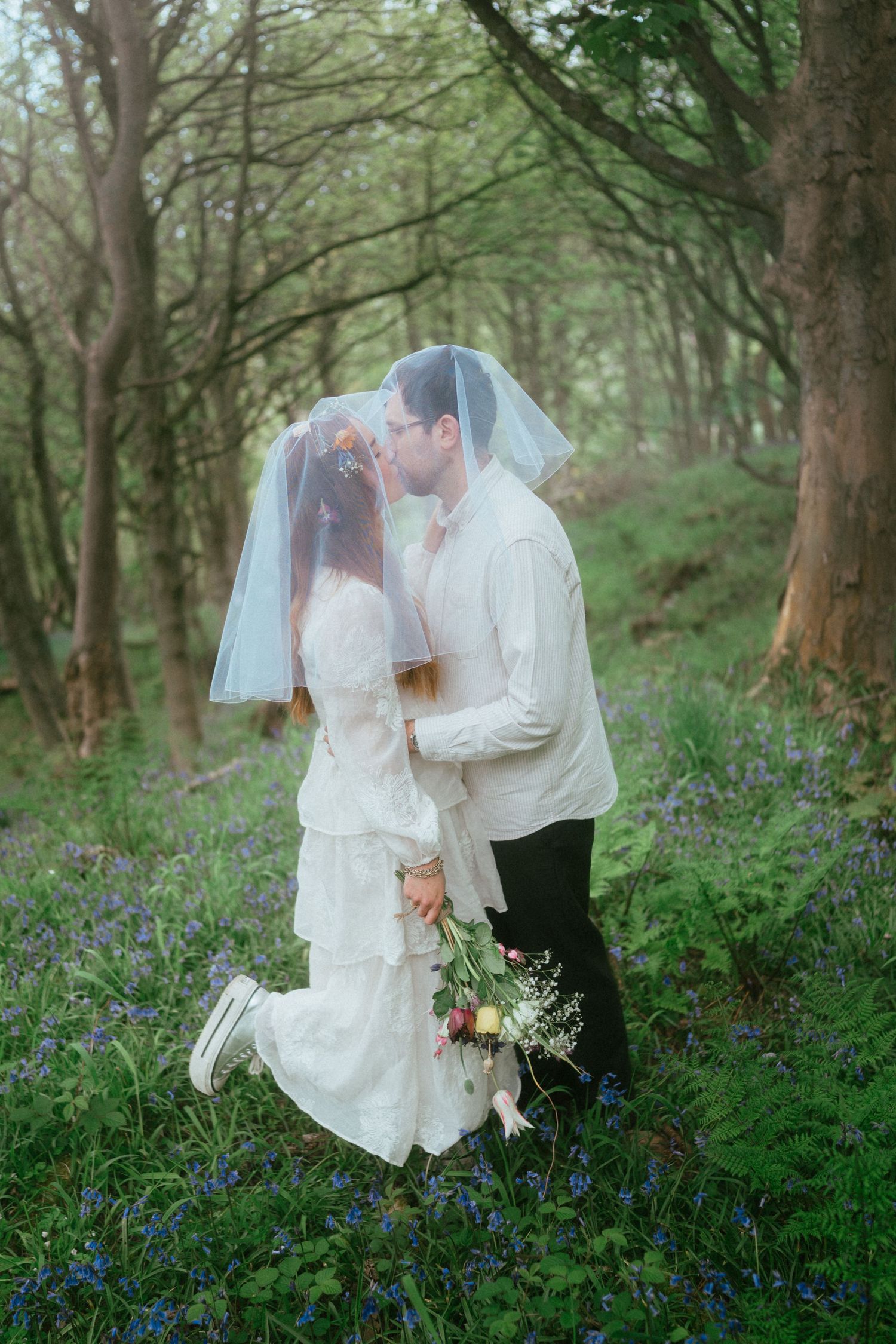 Bride and groom kissing under a veil in a bluebell woodland, bride holding a bouquet wearing a white lace dress.
